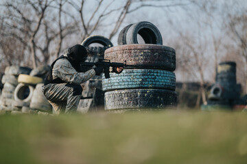 A paintball player crouches behind stacked tires, aiming a rifle in an outdoor field. Tactical action captures a moment during a game.