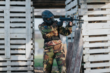 Fototapeta premium A masked airsoft player in camouflage holds a replica rifle behind a wooden bunker. The scene captures a tense moment in an outdoor airsoft game.