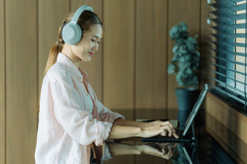 An Asian woman is taking notes while wearing headphones during an online class on a tablet or working remotely in a coffee shop.