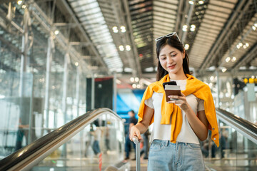 Fototapeta premium Happy young asian woman traveler holding boarding pass ticket, passport and smartphone at the airport terminal with her luggage and smiling at camera, cheerful tourist female having holiday trip.