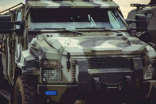 A horizontal close-up shot of the front section of a military armored vehicle featuring a multi-tone camouflage pattern and a reinforced bumper with a blue light.