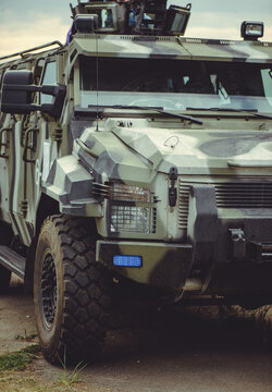 A vertical close-up shot of a rugged military armored vehicle with a camouflage pattern, featuring a blue tactical light on the bumper and reinforced headlights.
