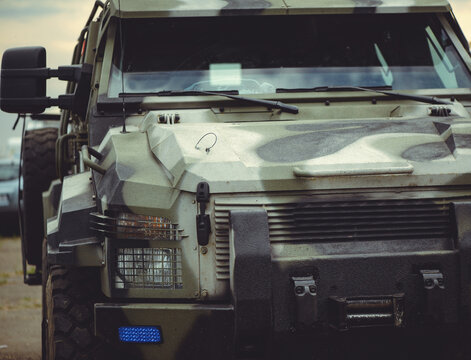 A close-up front view of a rugged military armored vehicle with a camouflage pattern, featuring reinforced headlights and a heavy-duty bumper.