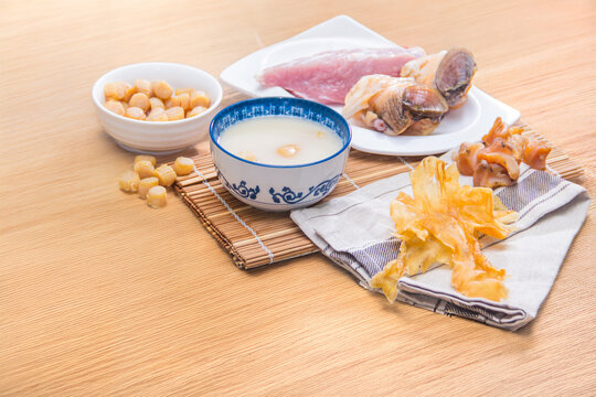 A ceramic bowl of white soup placed on a wooden table surrounded by dried scallops, pork, and mushrooms.