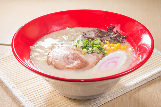 A red rim bowl holds creamy tonkotsu ramen with chashu, corn, scallion, wood ear, and a pink swirl fish cake. The rich broth and steam suggest classic Japanese comfort food.