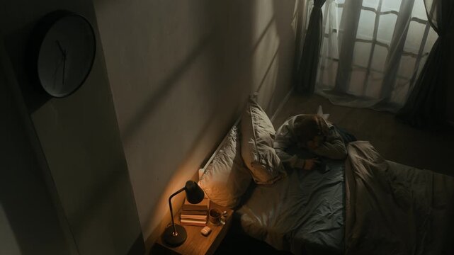 High angle lowkey shot of young woman with sleep deprivation sitting on floor by bed, suffering from fatigue and inability to sleep at night