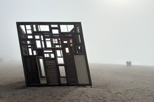 Abstract wooden frame installation standing on a foggy beach with distant figures in misty background. Liepaja, Veja Paveljons, 2026