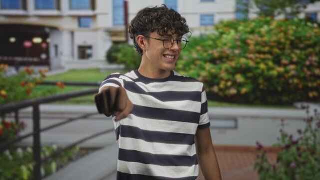 Teen boy points finger to camera, bare forearm visible, striped shirt in front of building; playful confidence.