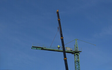 Construction worker in safety vest and hard hat overseeing the assembly of a tower crane jib, using a mobile crane boom against a clear blue sky background