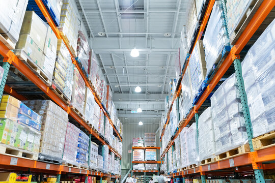 A wide interior photograph of a large warehouse-style wholesale shopping center featuring long aisles lined with tall pallet racks filled with bulk merchandise. The image captures the spacious retail 