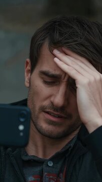 Vertical video: Reading text man rubbing brow at wall with phone wearing jacket and patterned shirt