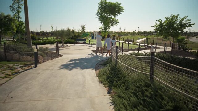 Back view of family of three dressed in white strolling along concrete path in beautifully landscaped park on bright, sunny day, enjoy quality time together during their holiday