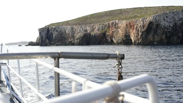 View of steep rocky cliffs along the coastline seen from the deck of a boat. The metal railing of the vessel is in the foreground over the calm sea.