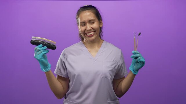 Woman dentist smiling widely holding tooth shade guide and dental mirror with gloved hands in purple studio; cheerful confidence.