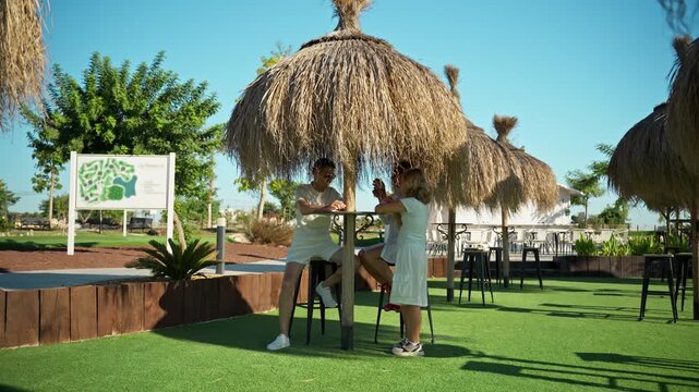 Tender moment of family under straw tiki umbrella on sunny day. Mother lovingly hugs daughter while father watches with joy. Family lifestyle, parenting, and heartwarming concept.