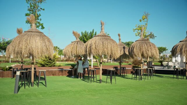 People enjoy sunny day at luxury resort, sit together in shade of straw tiki umbrella on lush green lawn, creating perfect scene of relaxation and leisure.Family lifestyle and happiness concept