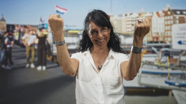 Woman with raised fists and bare arms smiling, wearing bracelets and necklace on a street by boats and buildings; joy victory resilience.