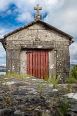 Fototapeta premium Traditional stone chapel, featuring a weathered crucifix on its roof and a rustic wooden door, embodying Galician heritage and a sense of enduring faith against a cloudy sky