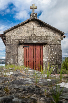 Traditional stone chapel, featuring a weathered crucifix on its roof and a rustic wooden door, embodying Galician heritage and a sense of enduring faith against a cloudy sky
