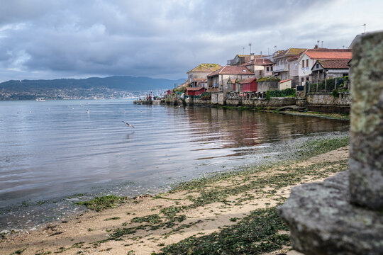 Combarro, Galicia, Spain, showing traditional stone houses with horreos and a sandy beach with seaweed, overlooking the calm Ria de Pontevedra on a cloudy day