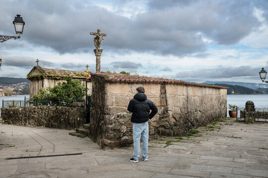 Man viewing the Ria de Pontevedra waterfront, gazing at a traditional Galician cruceiro and horreo, capturing the essence of the historic fishing village of Combarro under a cloudy sky