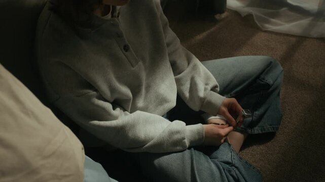 Handheld lowkey shot of sleepless Caucasian girl holding wristwatch in hands while sitting on floor by bed at dawn, lost in thought