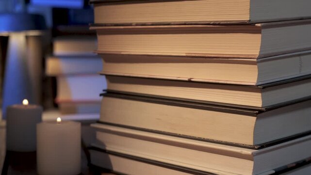 Stacked hardcover books beside candlelight creating cozy reading atmosphere, Quiet evening literature setting with warm illumination