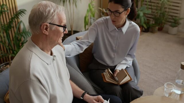 Handheld slowmo shot of empathetic middle-aged Caucasian female psychologist supporting senior male patient, gently touching him by shoulder during counseling session in modern office