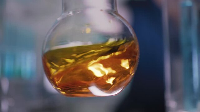 Female researcher preparing a new type of enriched bio fuel. Holding a glass flask with golden liquid close to the camera and slowly swirling the liquid. Selective focus