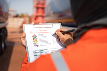 Fototapeta premium An engineer is checking on chemical hazardous material checklist to verify the safety condition with chemical road tanker as background. Industrial waorking scene, close-up and selective focus.