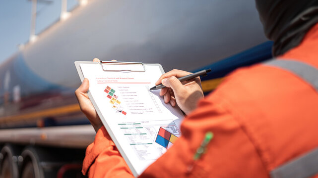 An engineer is checking on chemical hazardous material checklist to verify the safety condition with chemical road tanker as background. Industrial waorking scene, close-up and selective focus.