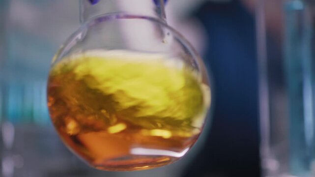 Female researcher preparing a new type of enriched fuel. Holding a glass flask with golden liquid close to the camera and swirling the liquid. Selective focus