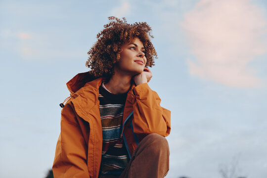 A smiling woman with curly hair wearing a rainbow sweater and orange jacket looks thoughtfully into the distance outdoors. The woman embodies a positive lifestyle and casual fashion.