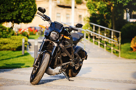 A horizontal shot of a sleek black electric motorcycle with orange turn signals, parked on a paved path with green trees and a building in the blurred background.