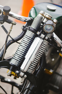 A vertical close-up shot of a classic custom motorcycle featuring a polished chrome springer fork suspension, a small round headlight, and a dark green fuel tank.