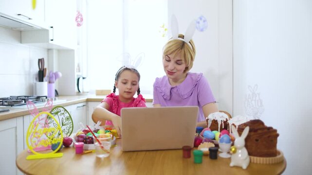 A mother and daughter wearing bunny ears celebrate Easter together online, surrounded by decorated eggs and traditional cakes, looking at camera.