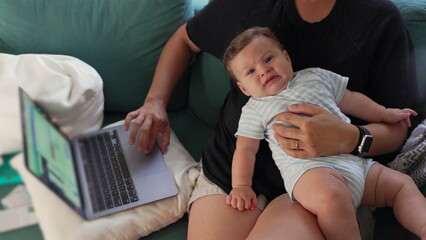 Mother holding baby’s hands while working on laptop, multitasking parent, balancing job and...