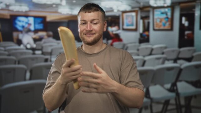 Man caucasian young heavyset holding a baguette and raising fist in building art gallery with rows of chairs and framed paintings, smiling; joy humor.