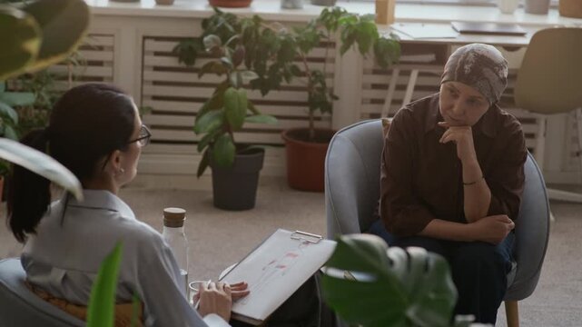 Over-the-shoulder slowmo shot of female psychologist analyzing human body diagram with colored marks during cancer counseling session with mature Asian female patient in modern office