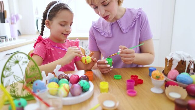 A mother and daughter joyfully paint colorful Easter eggs together at a wooden table, surrounded by craft supplies and traditional Easter cake