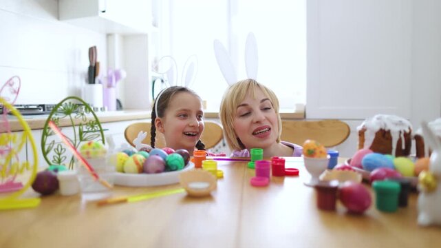 A mother and daughter wearing bunny ears peek over a table filled with colorful Easter eggs and painting supplies, ready for holiday activities