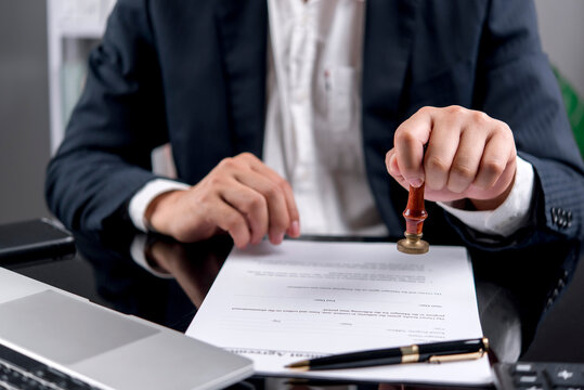 Businessman in navy suit stamping rubber seal on management agreement contract document on desk with laptop and pen. Legal approval, notary, authorization, corporate deal and certification concept.