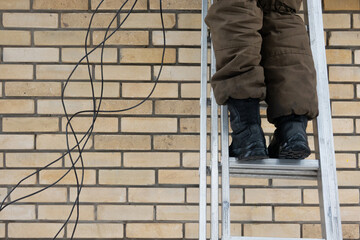 Worker's legs in brown pants and black boots on a metal ladder against a beige brick wall. Black cables hang nearby, suggesting construction or repair. Industrial feel with ample copy space © Анатолий Еремин