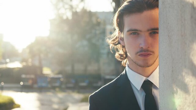 Young man in a stylish classic business suit with a tie, who peeks out from behind the wall and looks suspiciously into the distance  with sunlight during sunset.