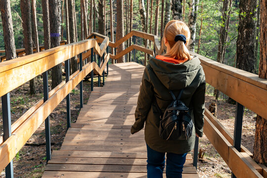Young Woman Walking On Wooden Boardwalk Through Pine Forest With Backpack And Jacket