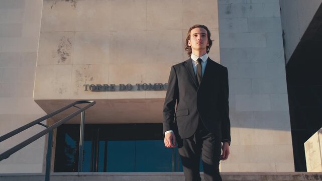 Young confident man in a stylish classic business suit with a tie walks down the steps and stares intently at the camera against the background of a bright building during sunset. Slow motion.