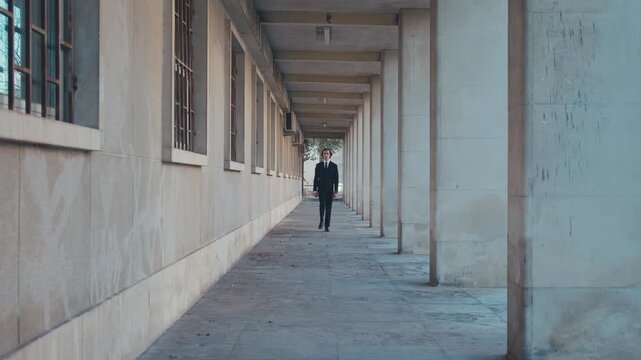 A stylish young man with medium-length hair in an elegant classic business suit and tie walks in a deserted city along large columns. Slow motion.