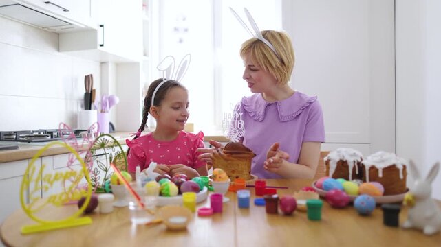 A mother and daughter wearing bunny ears prepare for Easter by decorating cakes and painting eggs in a bright kitchen setting
