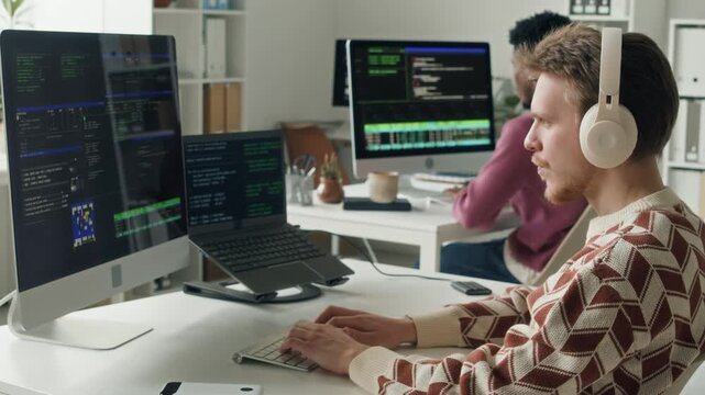 Side view of young adult male IT engineer wearing headset typing code on desktop computer at desk with laptop while troubleshooting script next to colleagues desk in open plan office