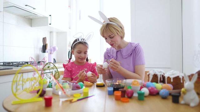 A mother and daughter are happily decorating white eggs with wax for Easter, creating colorful designs together at a wooden table